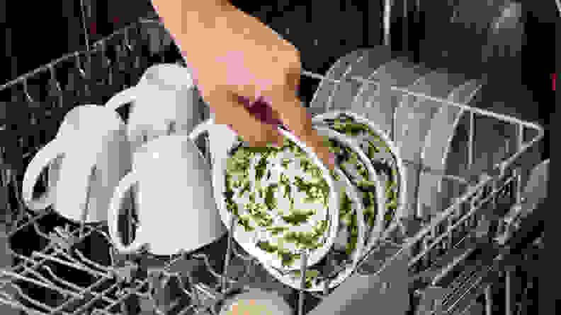 A hand loads a dirty dish into the upper rack of a dishwasher.