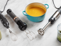 Three immersion blenders on a marble surface alongside blended soup, pesto, and a smoothie