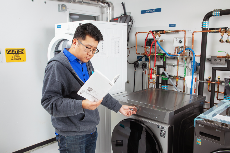 An Asian man in a gray sweatshirt reads a white manual and turns a dial on a washing machine