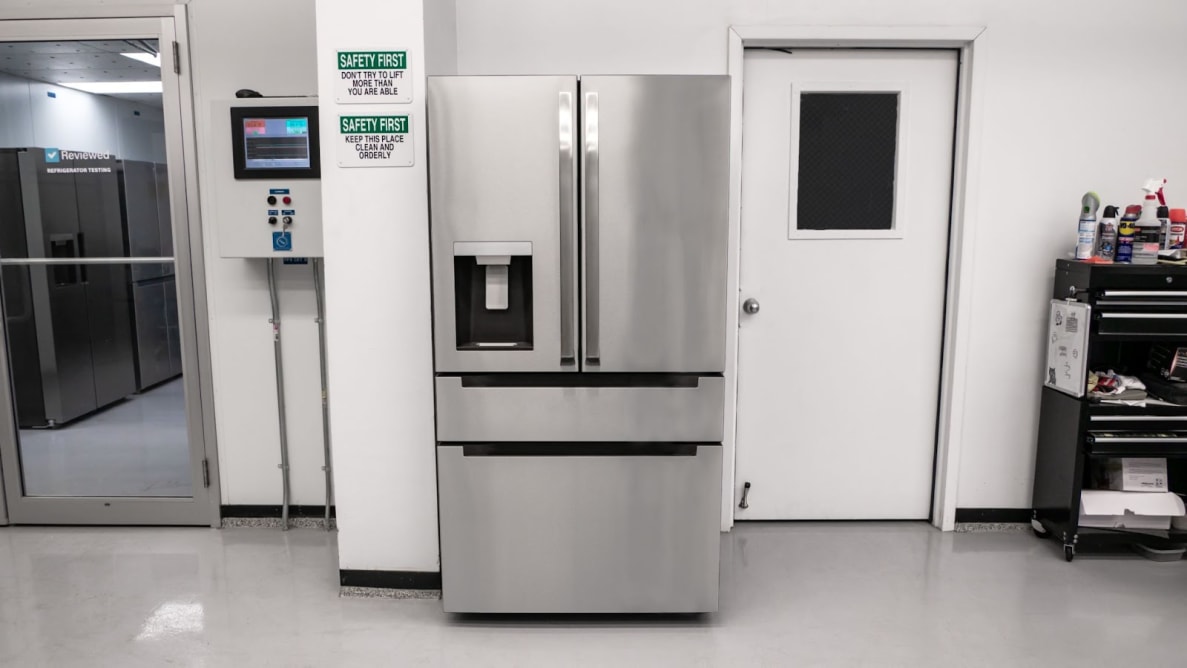 A stainless-steel refrigerator with exterior dispenser stands in a white lab environment next to a white door with a black window.