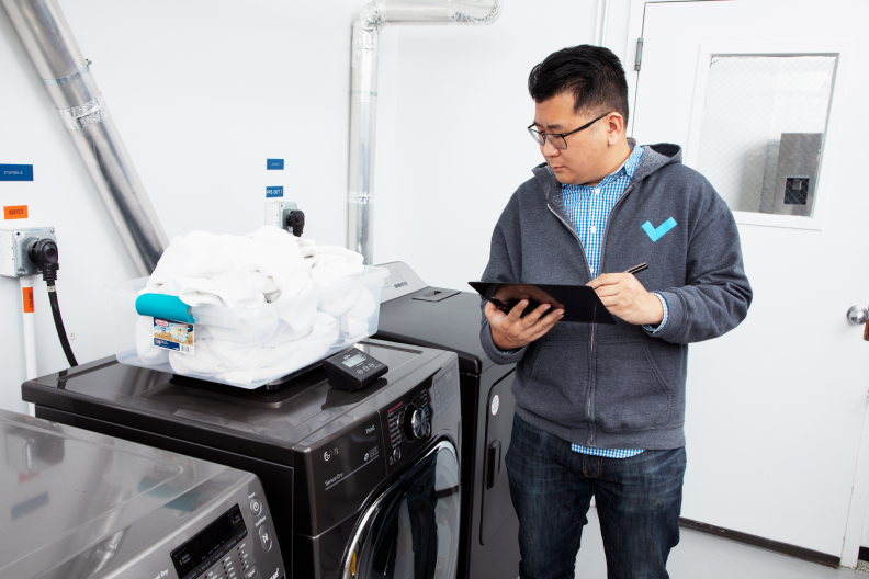 Person stares at clear storage container filled with white shirts on top of scale that sits on black dryer while writing on clipboard in front of white wall indoors.